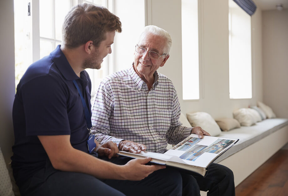 Elderly man and young caregiver looking at a photo album together in a bright room.