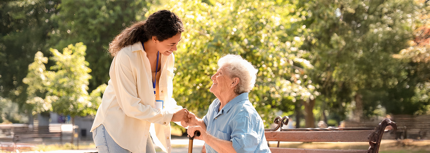 Senior woman sits on an outdoor bench smiling up at younger female caretaker